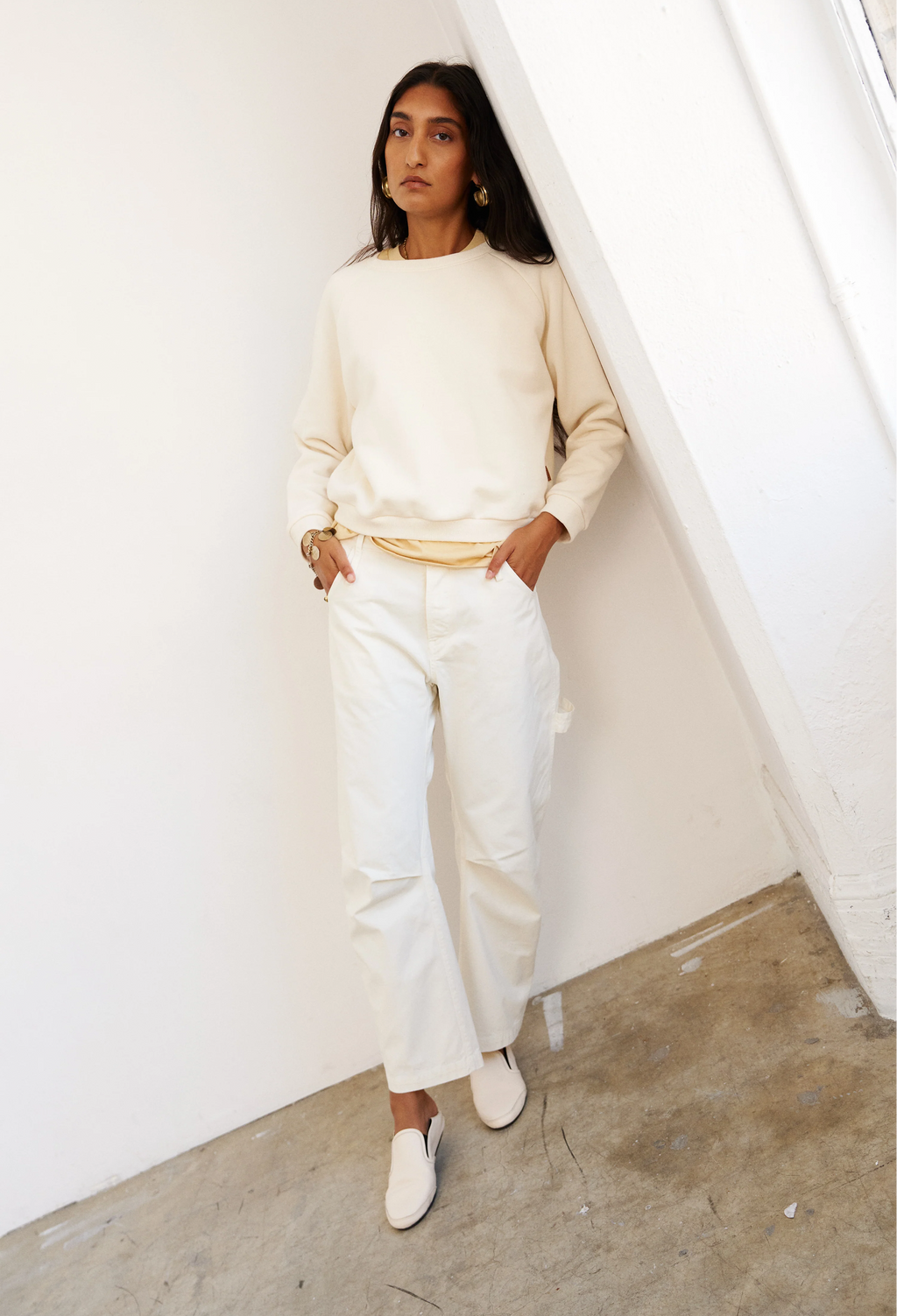 Woman wearing a cream sweater and white pants standing against a white wall.
