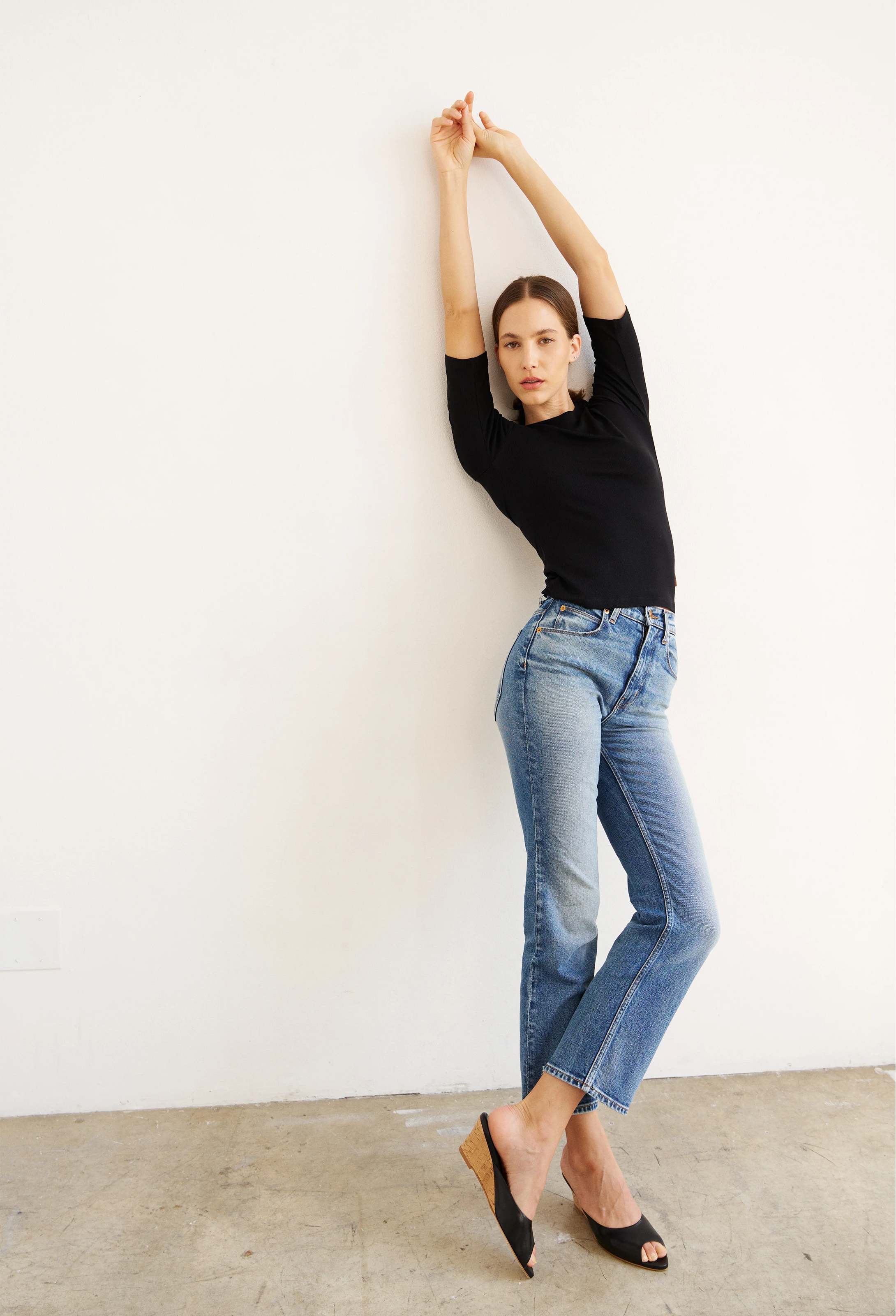 Woman wearing a black top and blue jeans against a white wall.