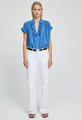 Woman wearing a blue blouse with ruffled details and white pants on a light gray background