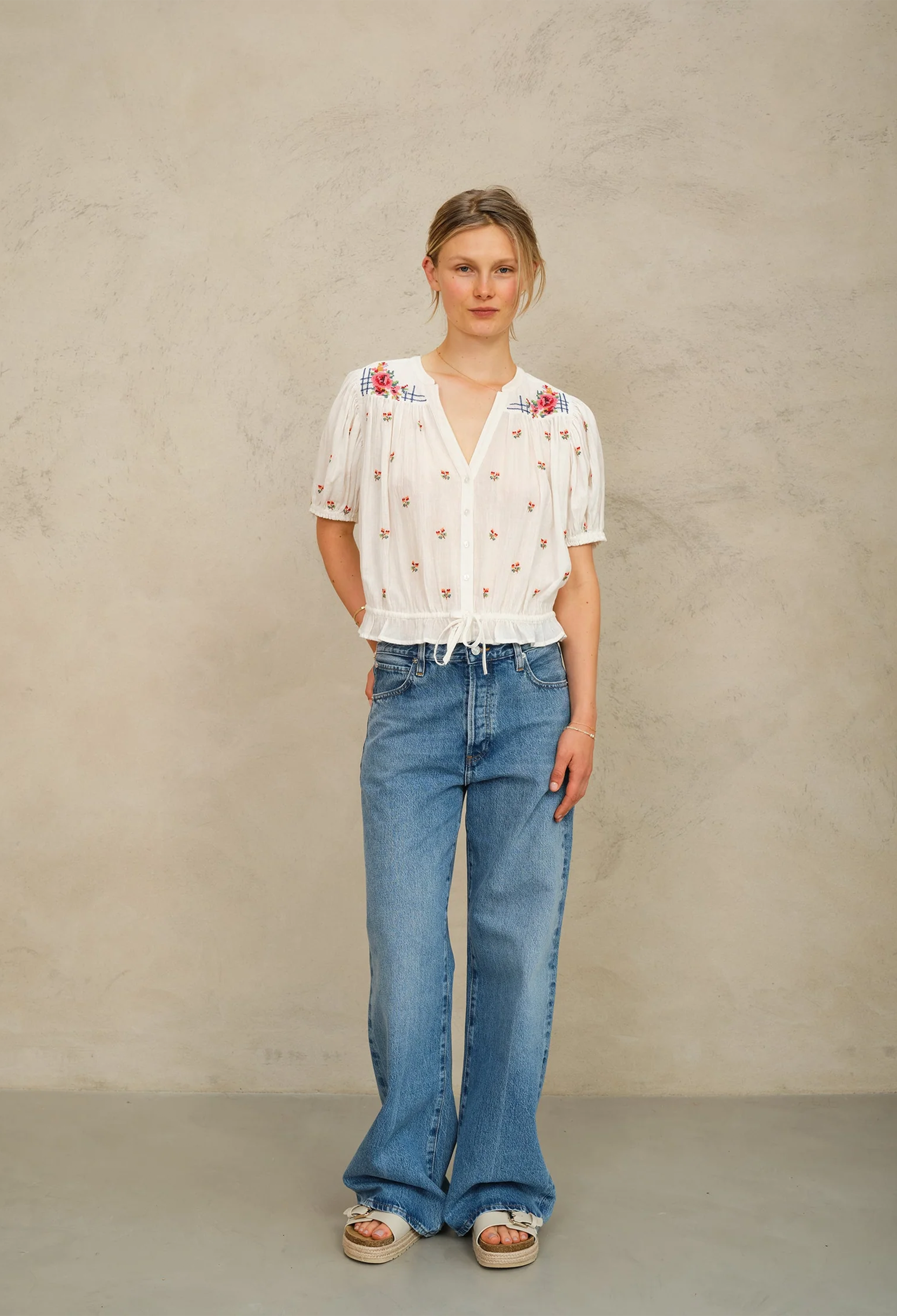 Woman wearing a white blouse with floral patterns and blue jeans against a plain background