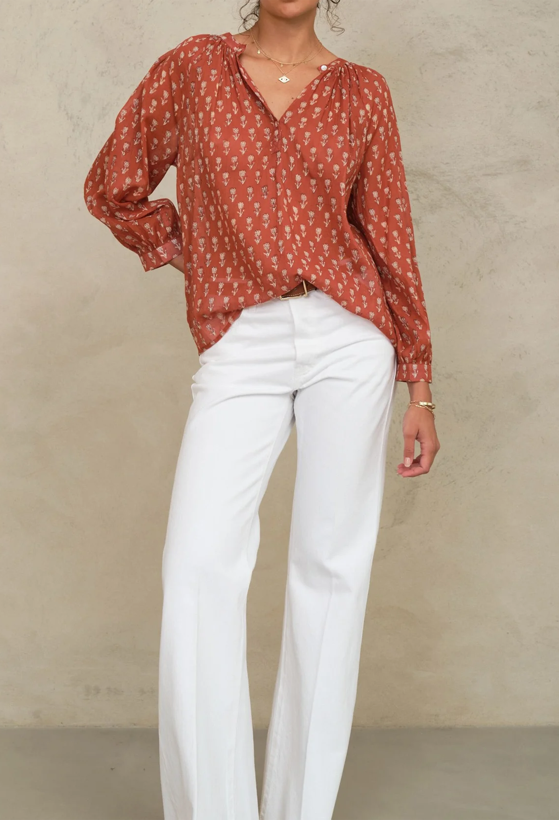Person wearing a rust-colored patterned blouse and white pants against a beige background