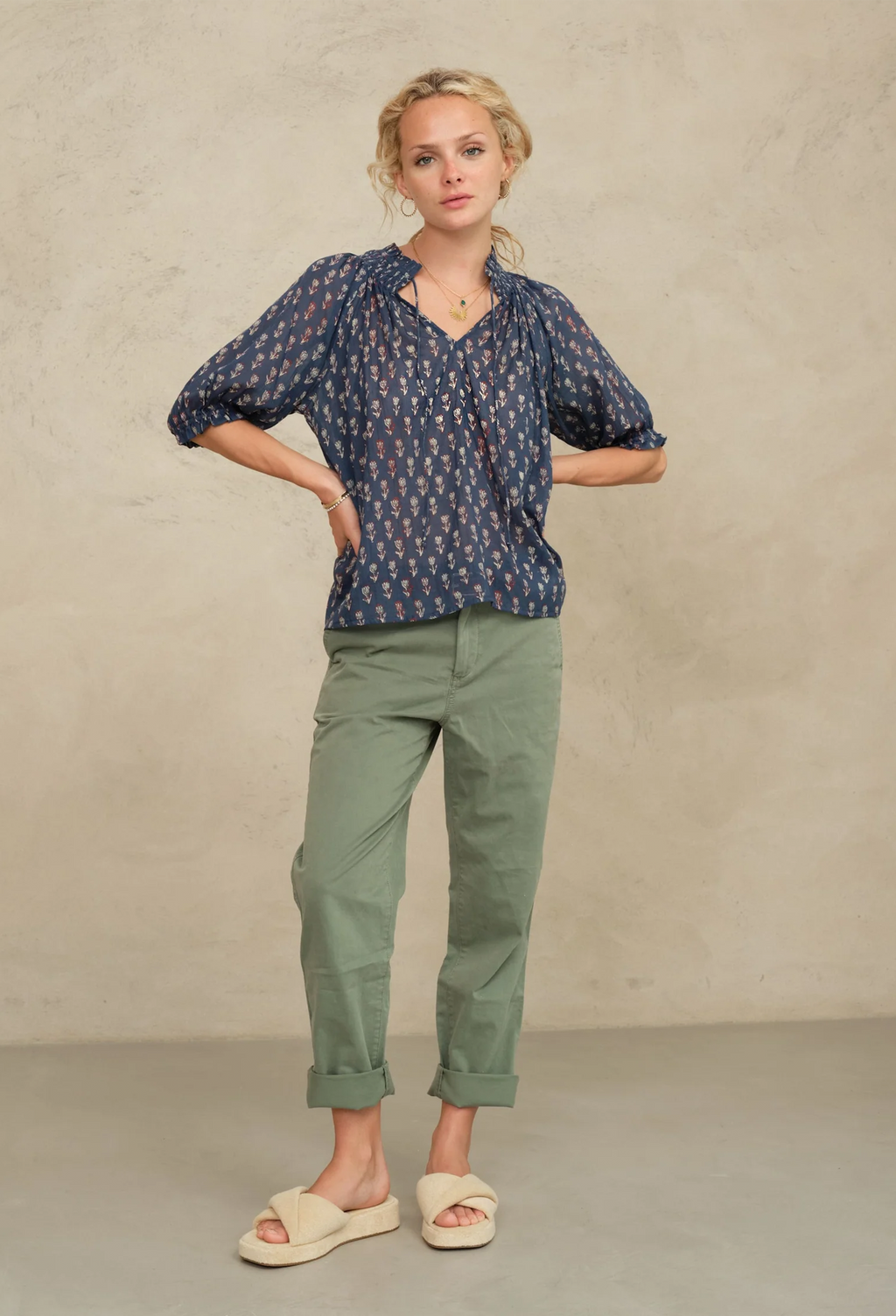 Woman wearing a blue patterned blouse and green pants against a beige background