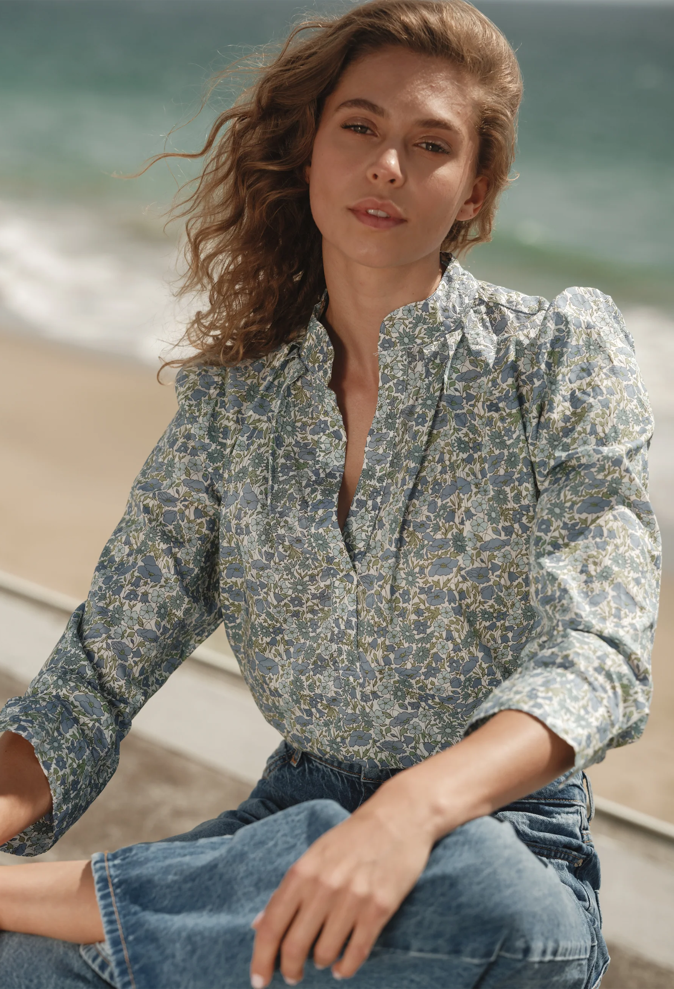 Woman sitting on a beach wearing a floral blouse and jeans.