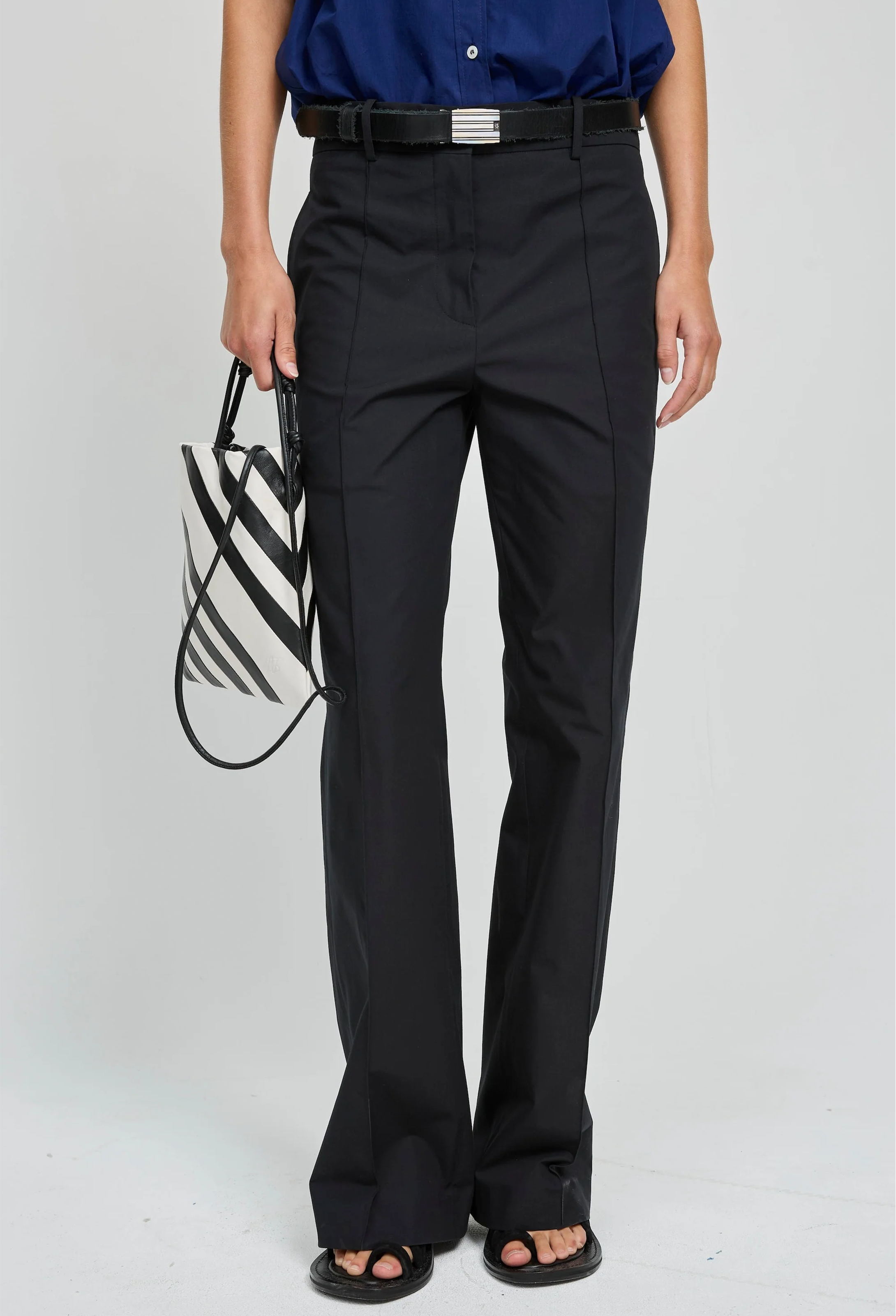 Person wearing black pants and a blue shirt, holding a black and white striped bag on a light gray background
