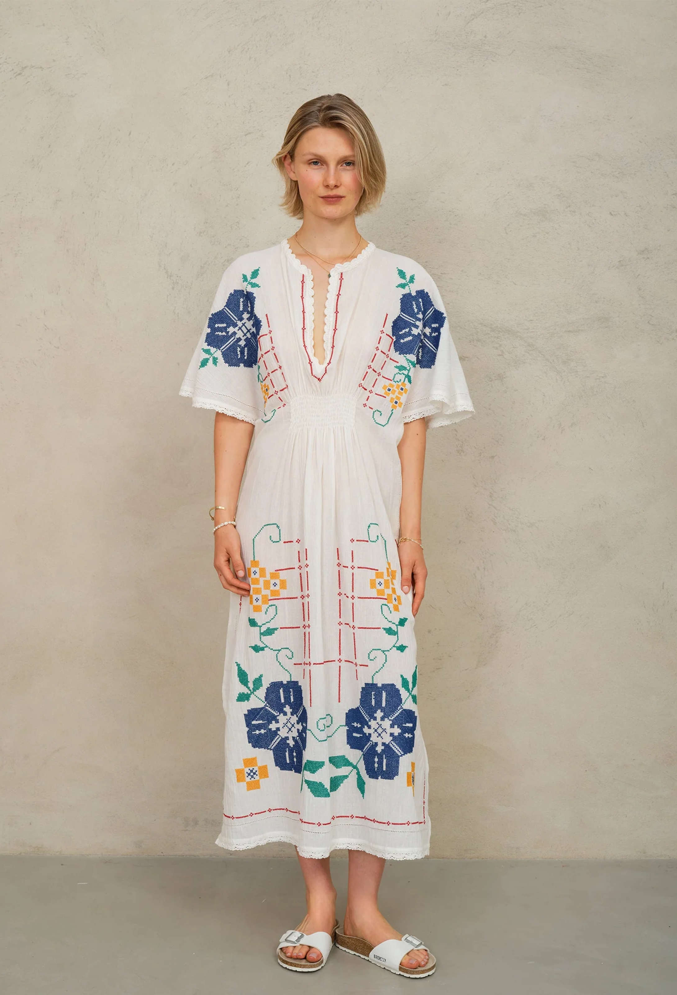 Woman wearing a white dress with colorful floral embroidery against a beige background