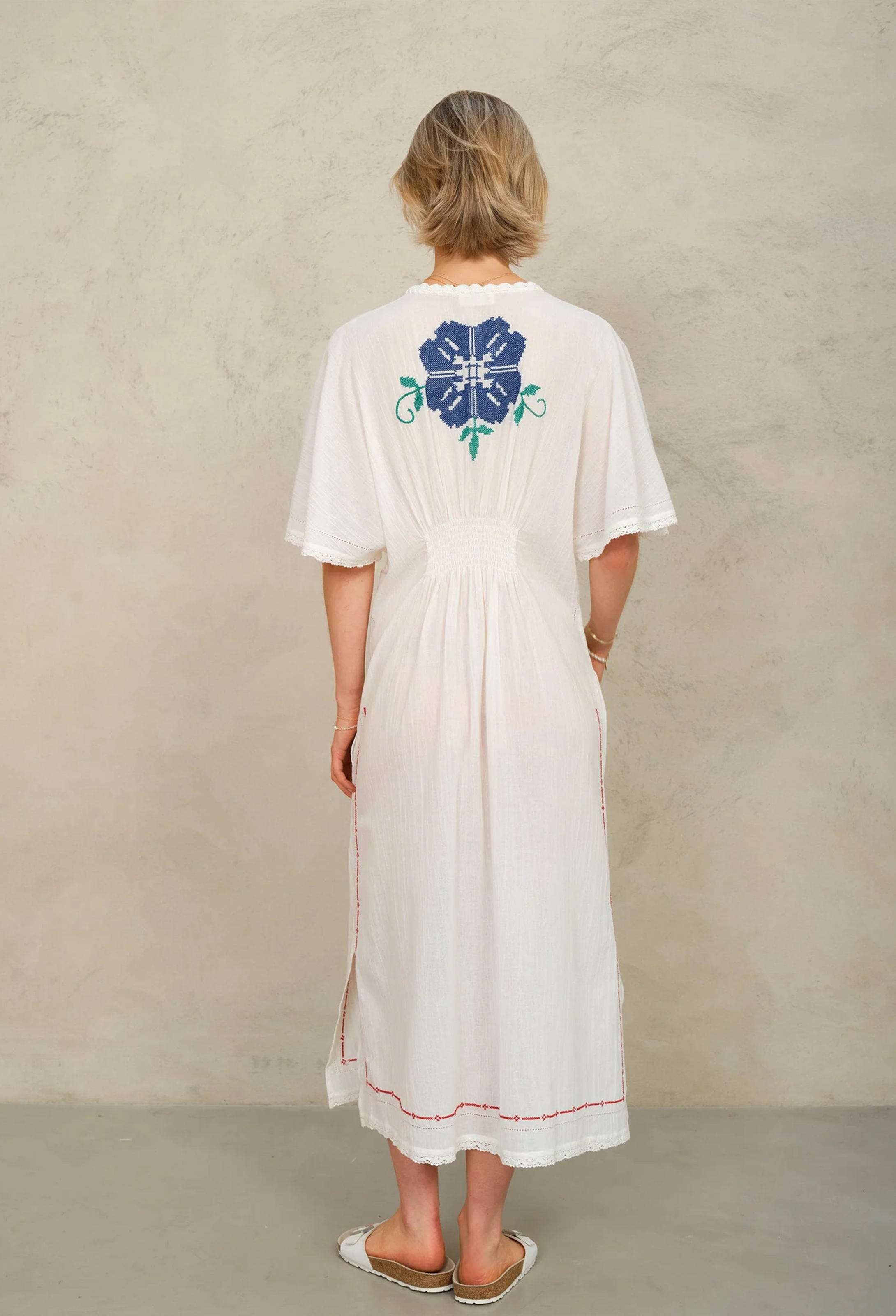 Woman wearing a white dress with a blue floral design on the back against a beige wall.