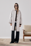 Person wearing a long beige coat in a minimalistic room with a chair.