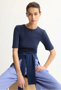 Woman wearing a navy top and blue striped pants sitting on a wooden stool against a white background