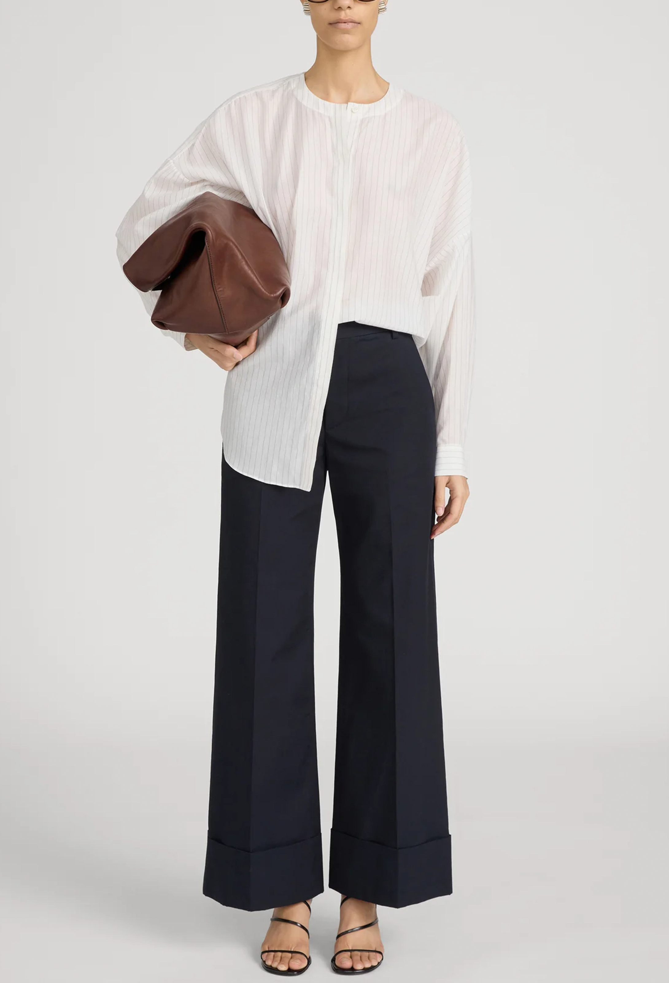 Person wearing a white blouse and navy pants holding a brown leather clutch against a white background