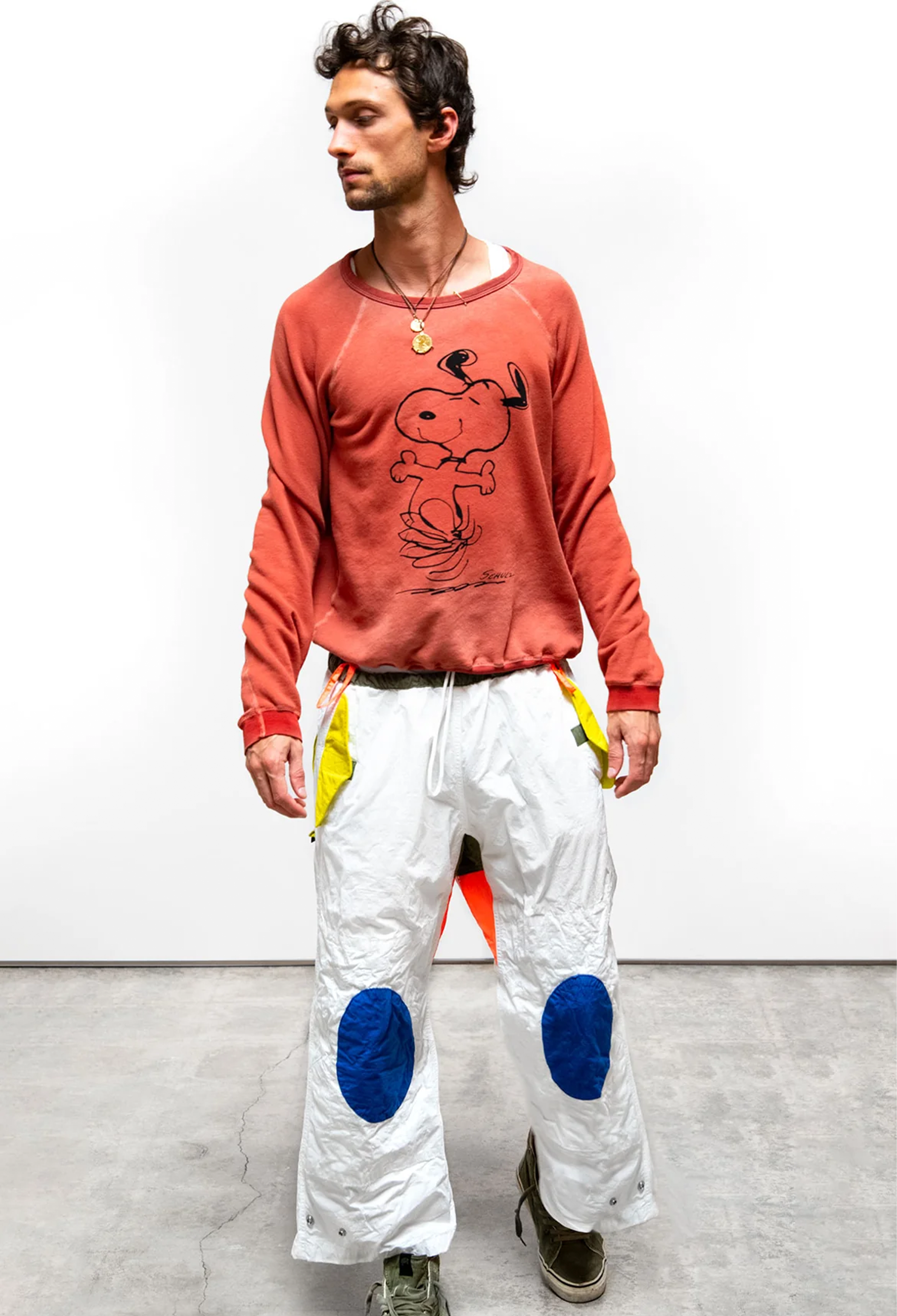 Person wearing a red sweatshirt with a graphic design and white pants with blue patches on a plain background