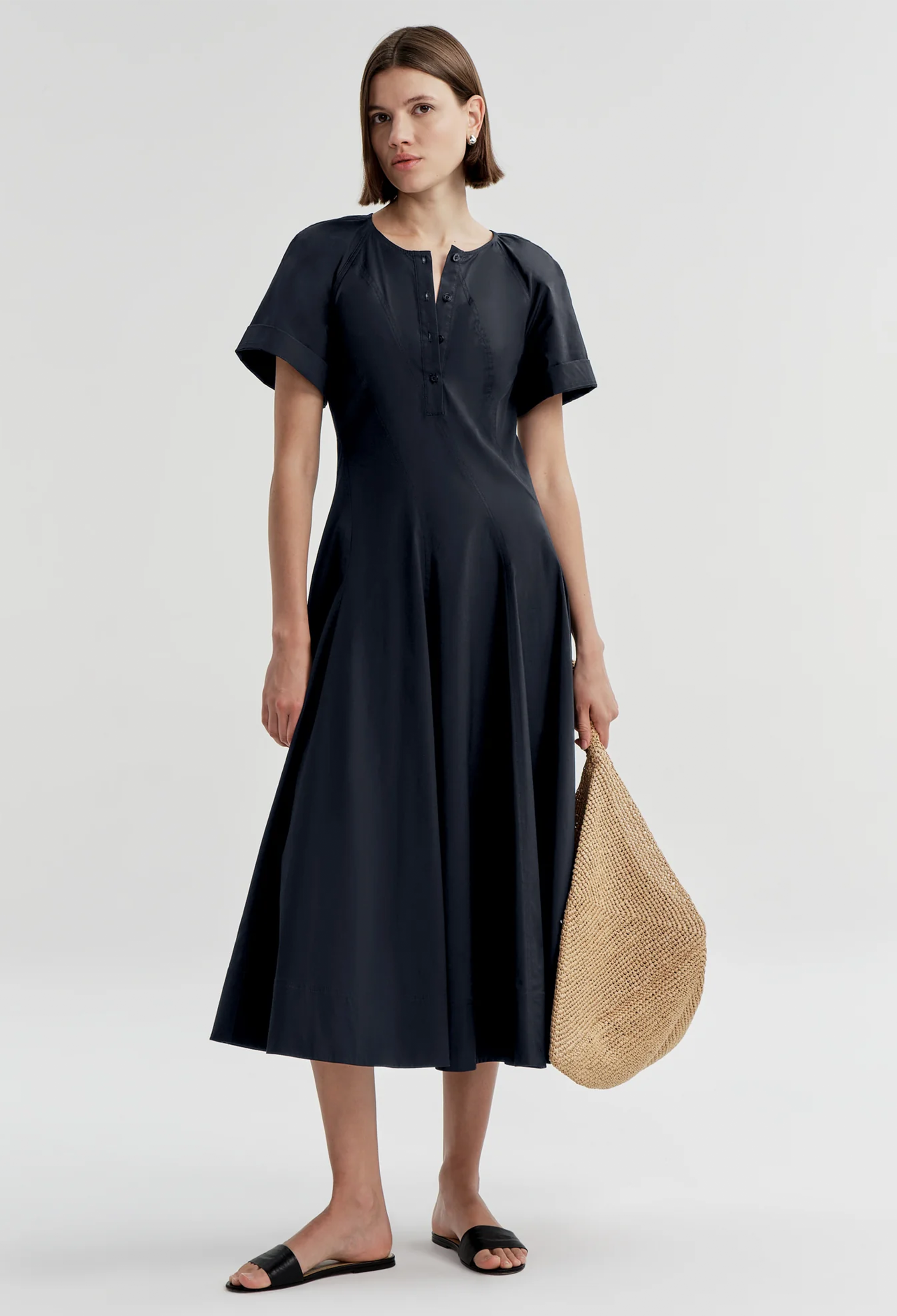 Woman wearing a navy dress holding a straw bag on a white background