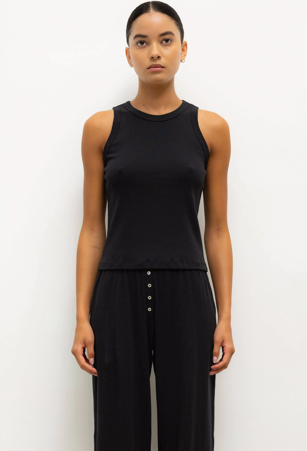 Person wearing a black sleeveless top and black pants with button details on a white background