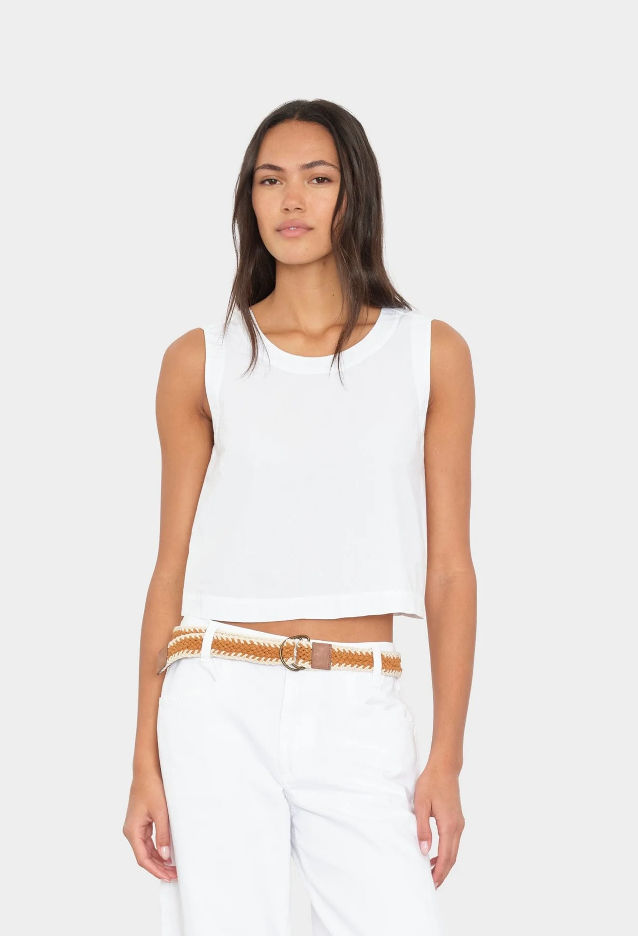 Woman wearing a white sleeveless top and white pants with a brown belt on a white background