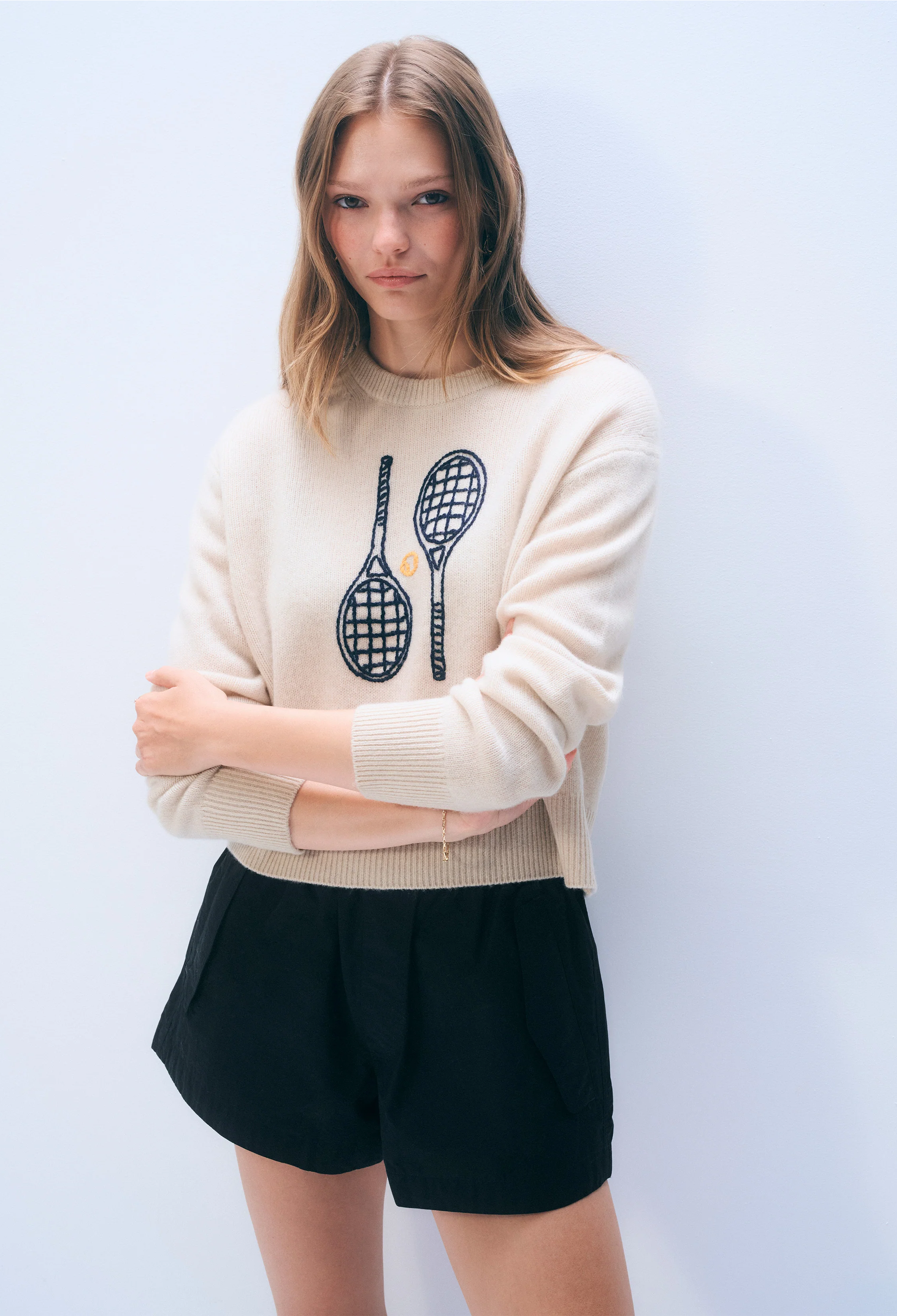 Woman wearing a cream sweater with tennis racket design and black shorts against a light blue background