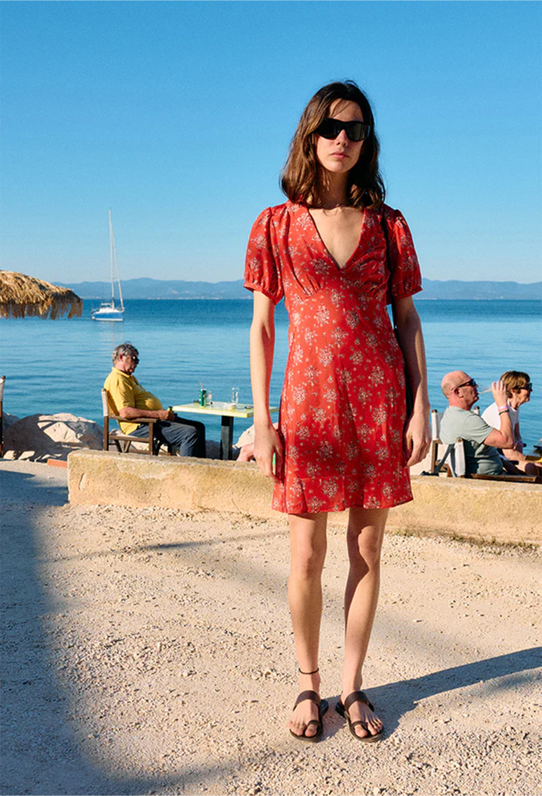 Woman in a red dress standing by the sea with people in the background