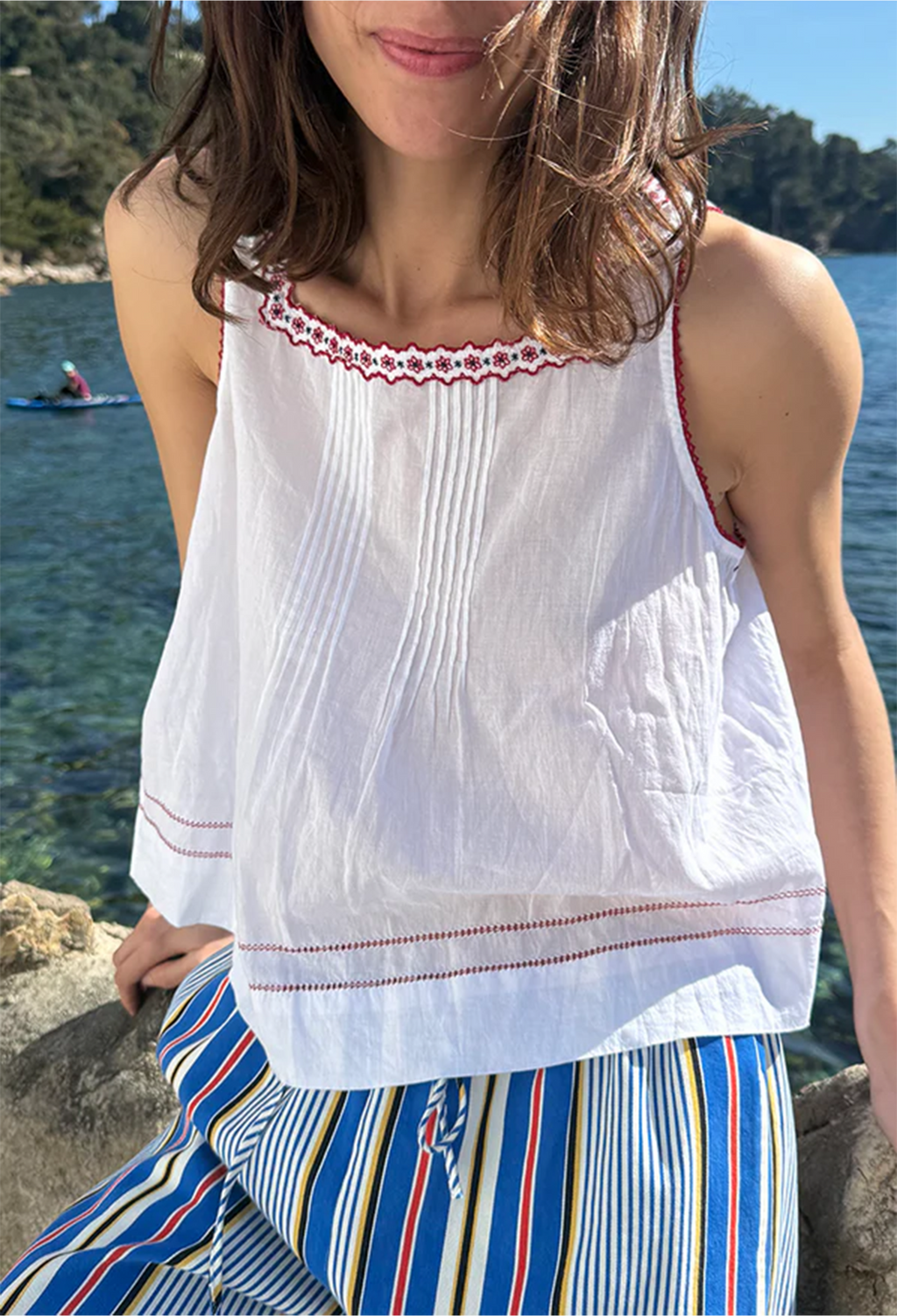 Woman wearing a white top with red embroidery and striped pants by a body of water.