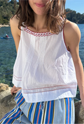 Woman wearing a white top with red embroidery and striped pants by a body of water.