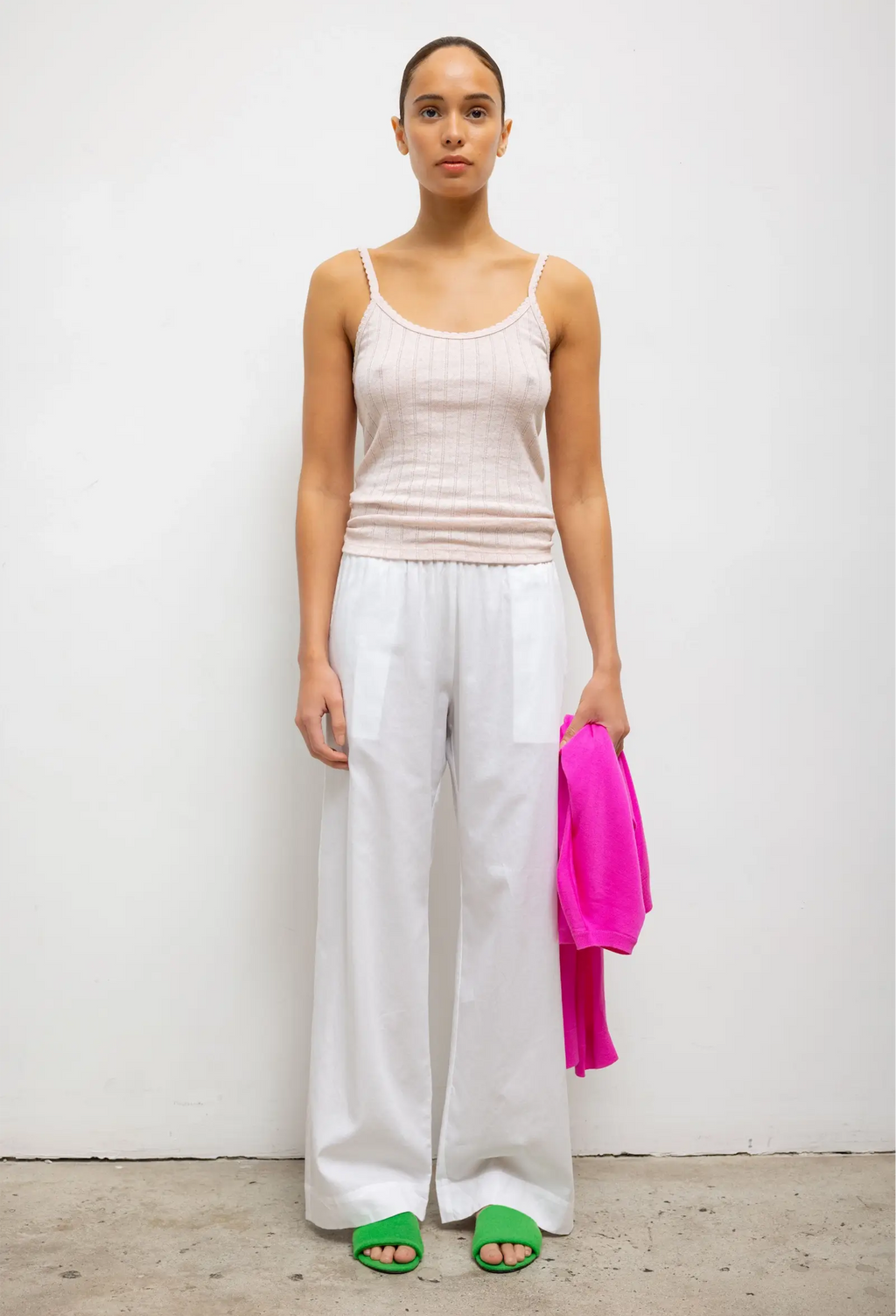 Woman wearing a beige tank top and white pants holding a pink jacket against a white background