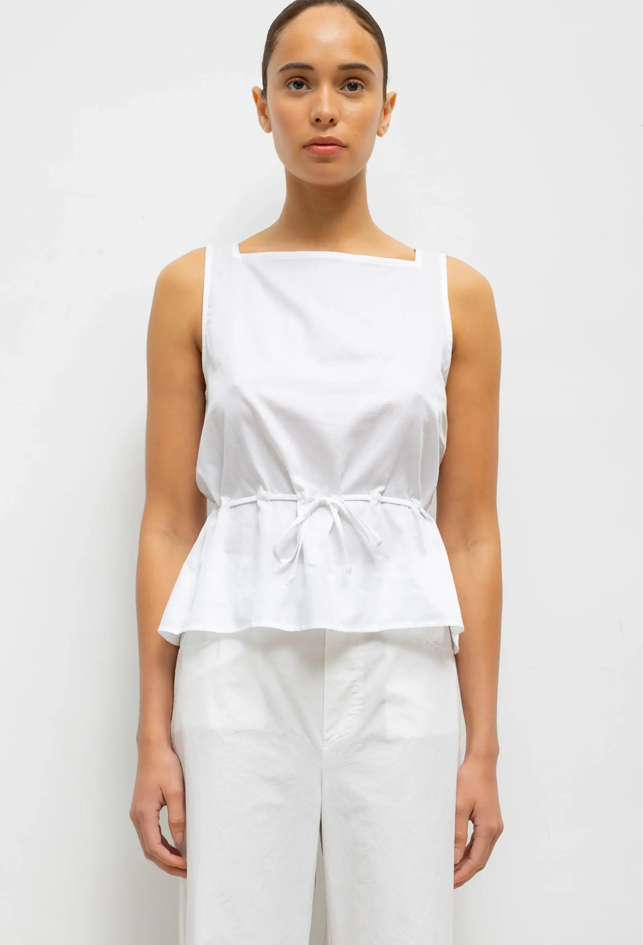 Woman wearing a white sleeveless top and pants against a plain background