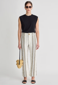 Woman wearing a black sleeveless top and striped pants, holding a woven bag on a light gray background