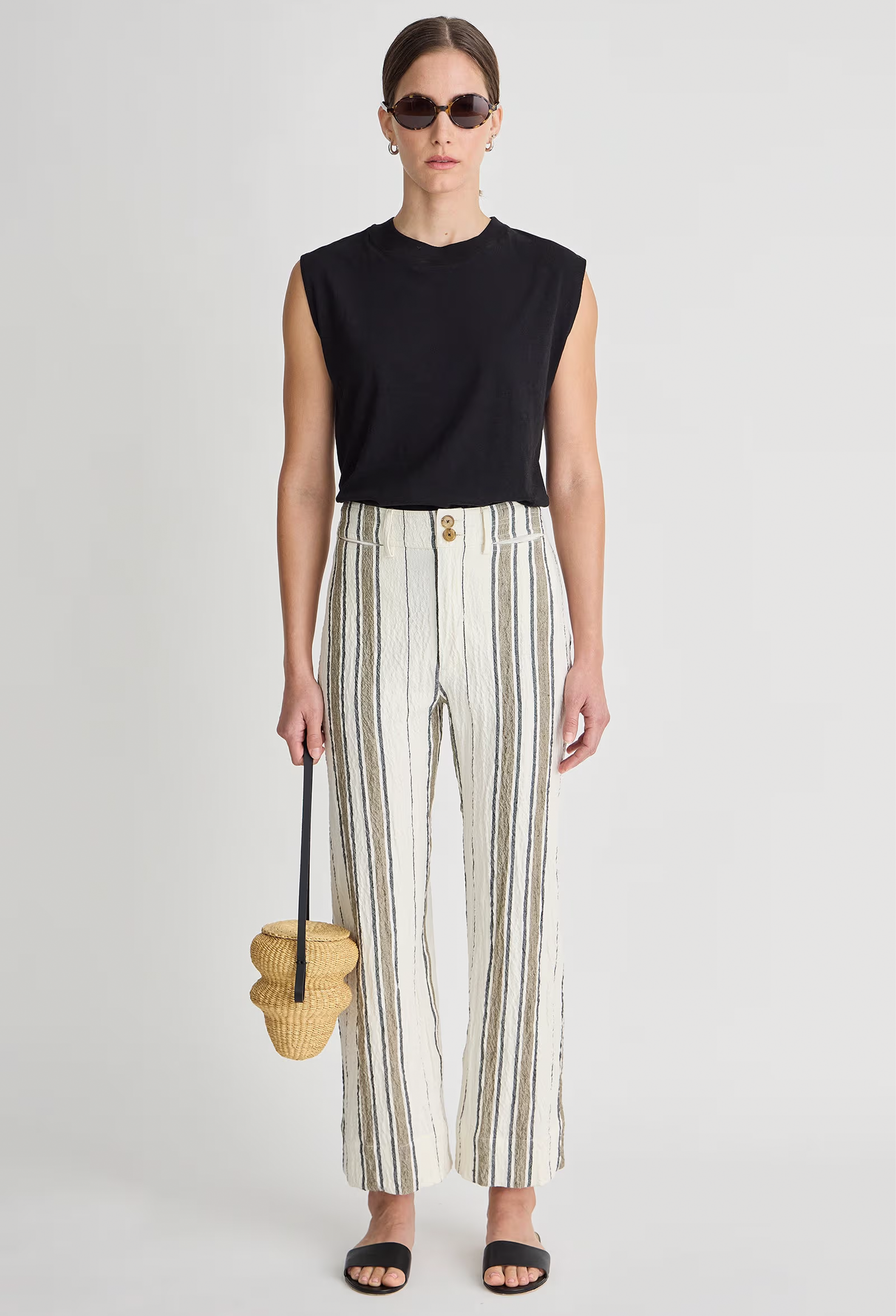 Woman wearing a black sleeveless top and striped pants, holding a woven bag on a light gray background