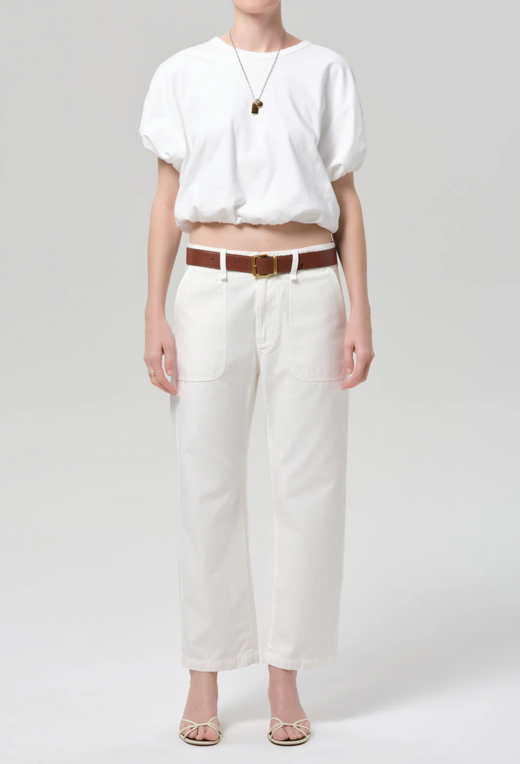 Person wearing a white blouse and white pants with a brown belt on a light gray background