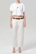 Person wearing a white blouse and white pants with a brown belt on a light gray background