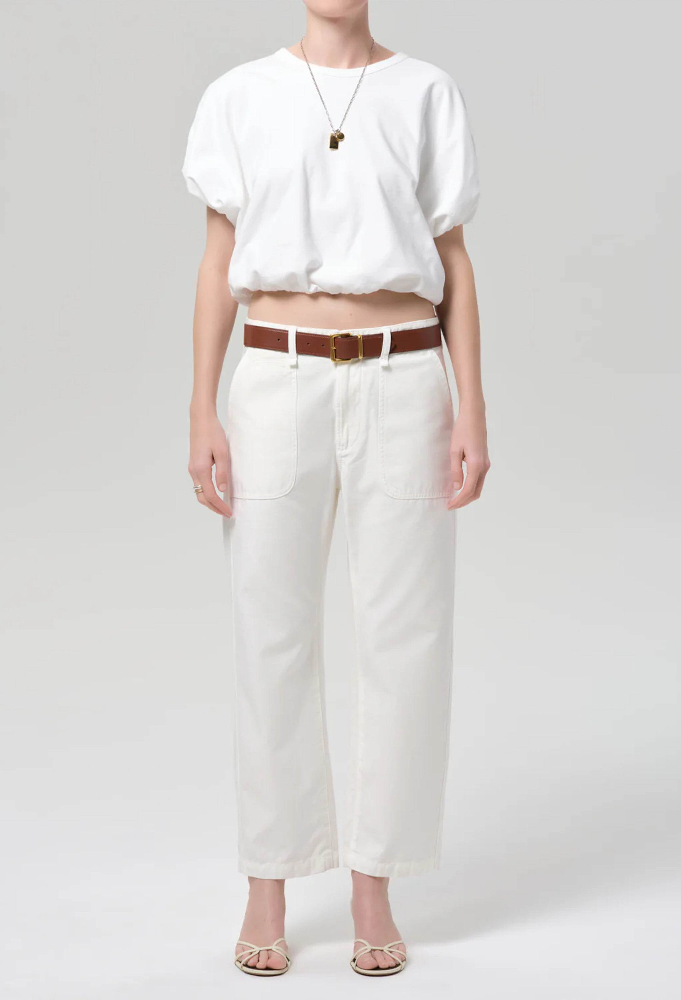 Person wearing a white blouse and white pants with a brown belt on a light gray background