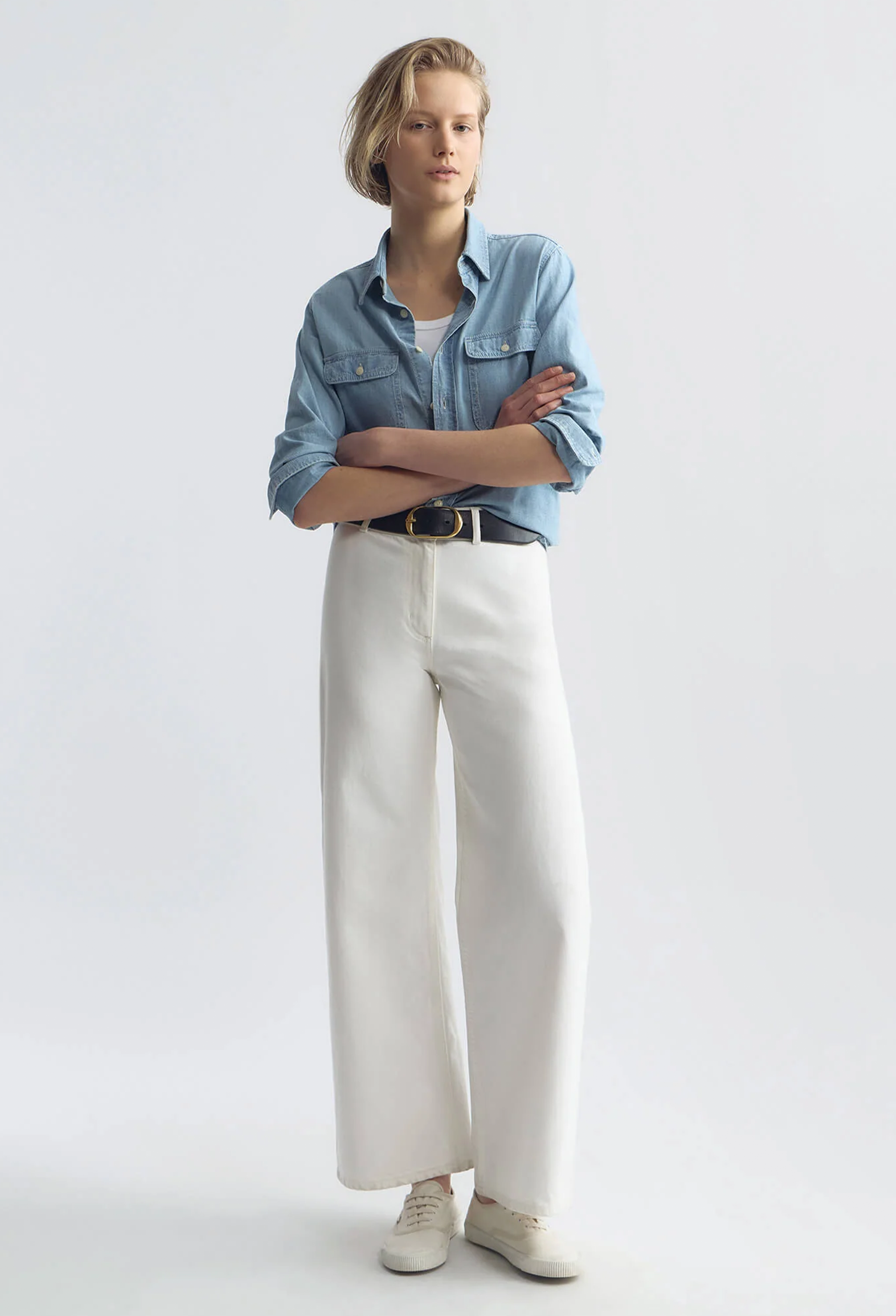 Person wearing a blue denim shirt, white pants, and white sneakers on a light gray background