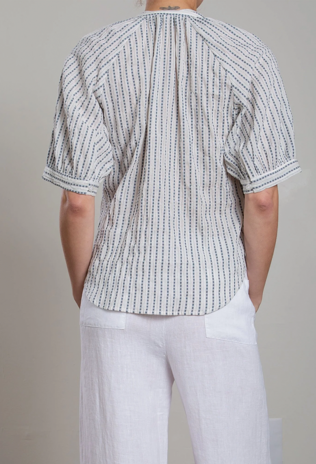 Person wearing a short-sleeve, patterned shirt and white pants against a neutral background