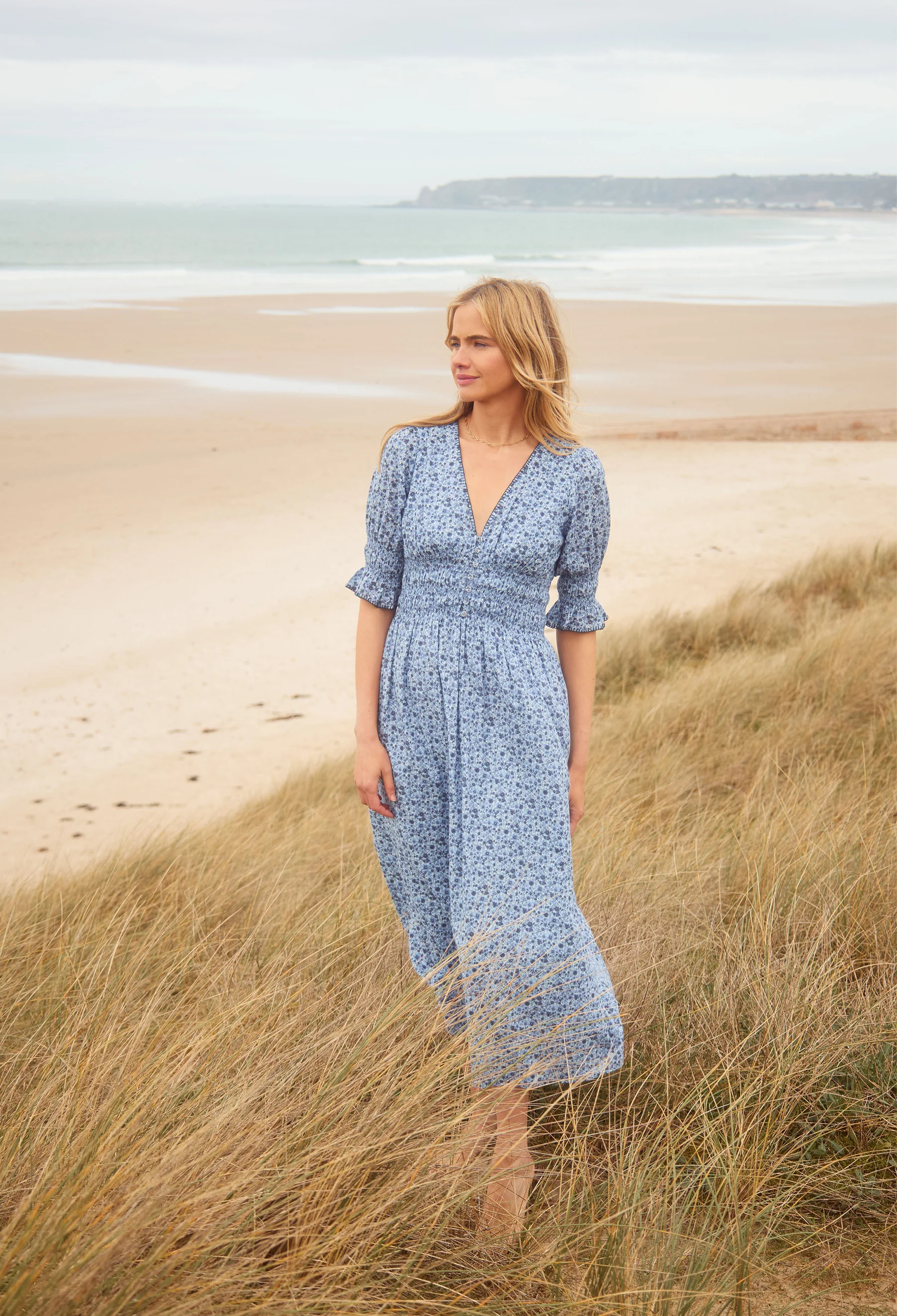 Woman in a blue dress standing on a beach with grass in the foreground