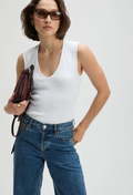 Woman wearing a white sleeveless top and blue jeans, holding a brown leather bag against a white background