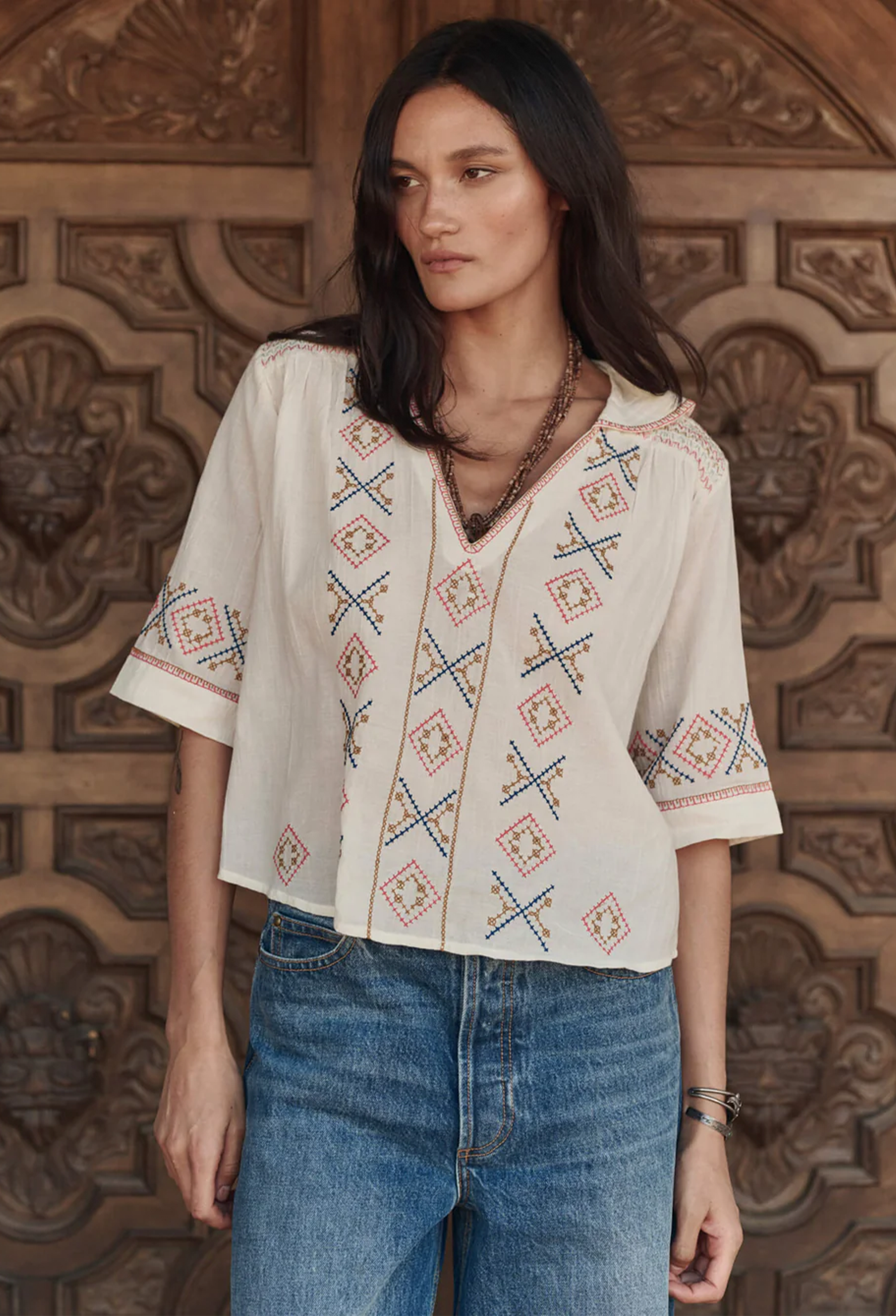 Woman wearing a patterned blouse and jeans standing against an ornate wooden wall.