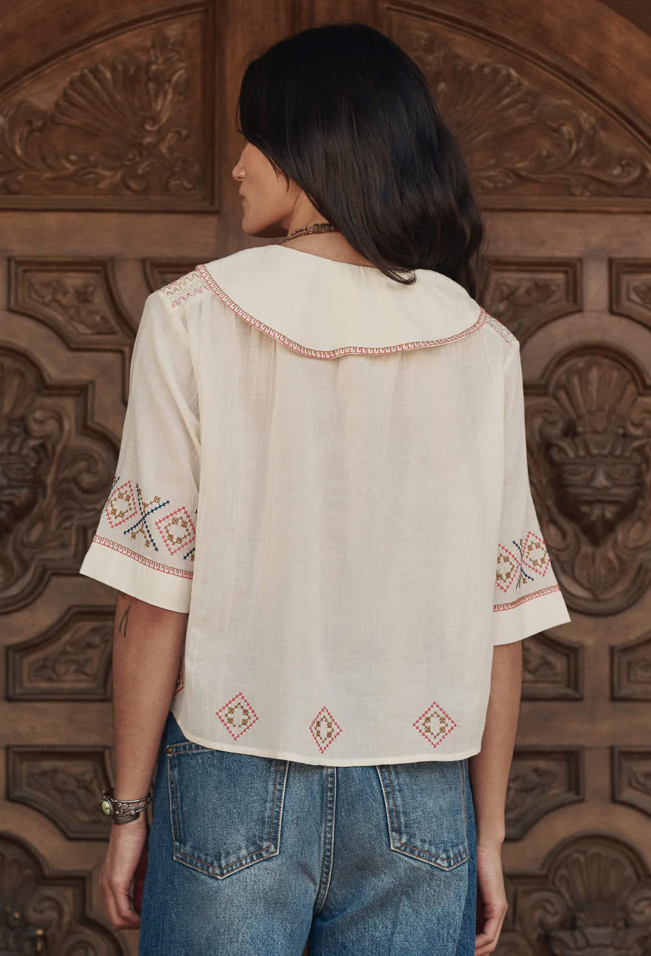 Person wearing a white embroidered blouse with blue jeans standing in front of a wooden carved door.