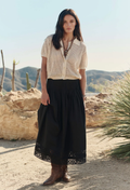 Woman in a white blouse and black skirt standing in a desert landscape with mountains in the background.