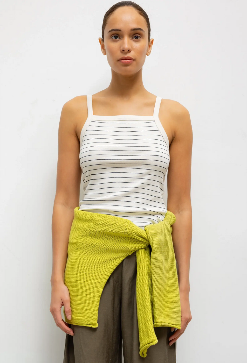 Woman wearing a white striped tank top and brown pants with a green wrap, standing against a white background