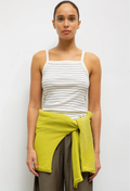 Woman wearing a white striped tank top and brown pants with a green wrap, standing against a white background