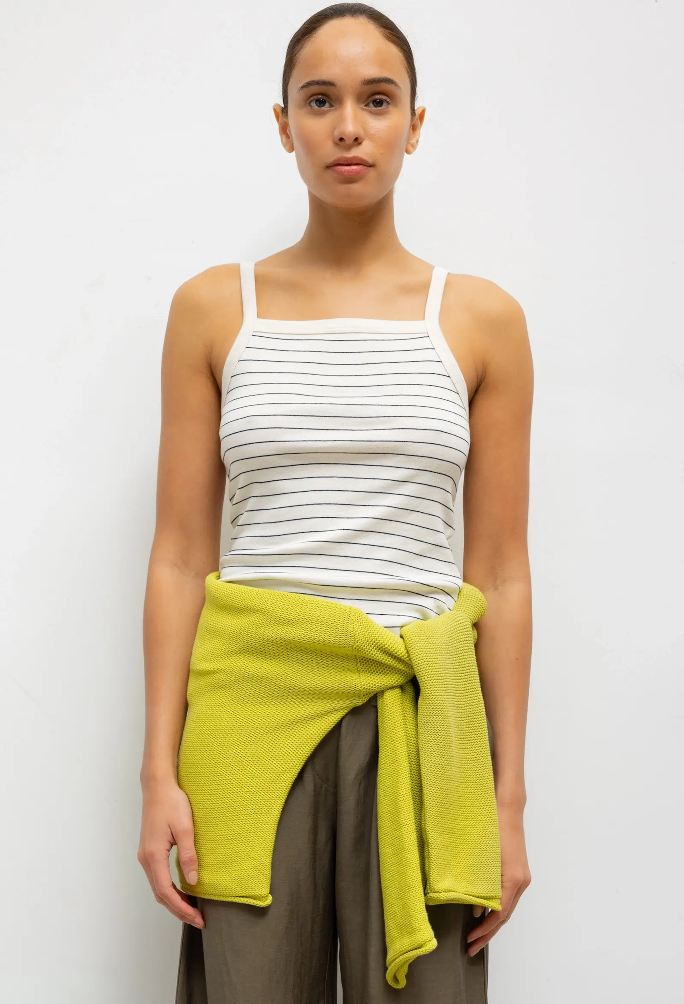 Woman wearing a white striped tank top and brown pants with a green wrap, standing against a white background