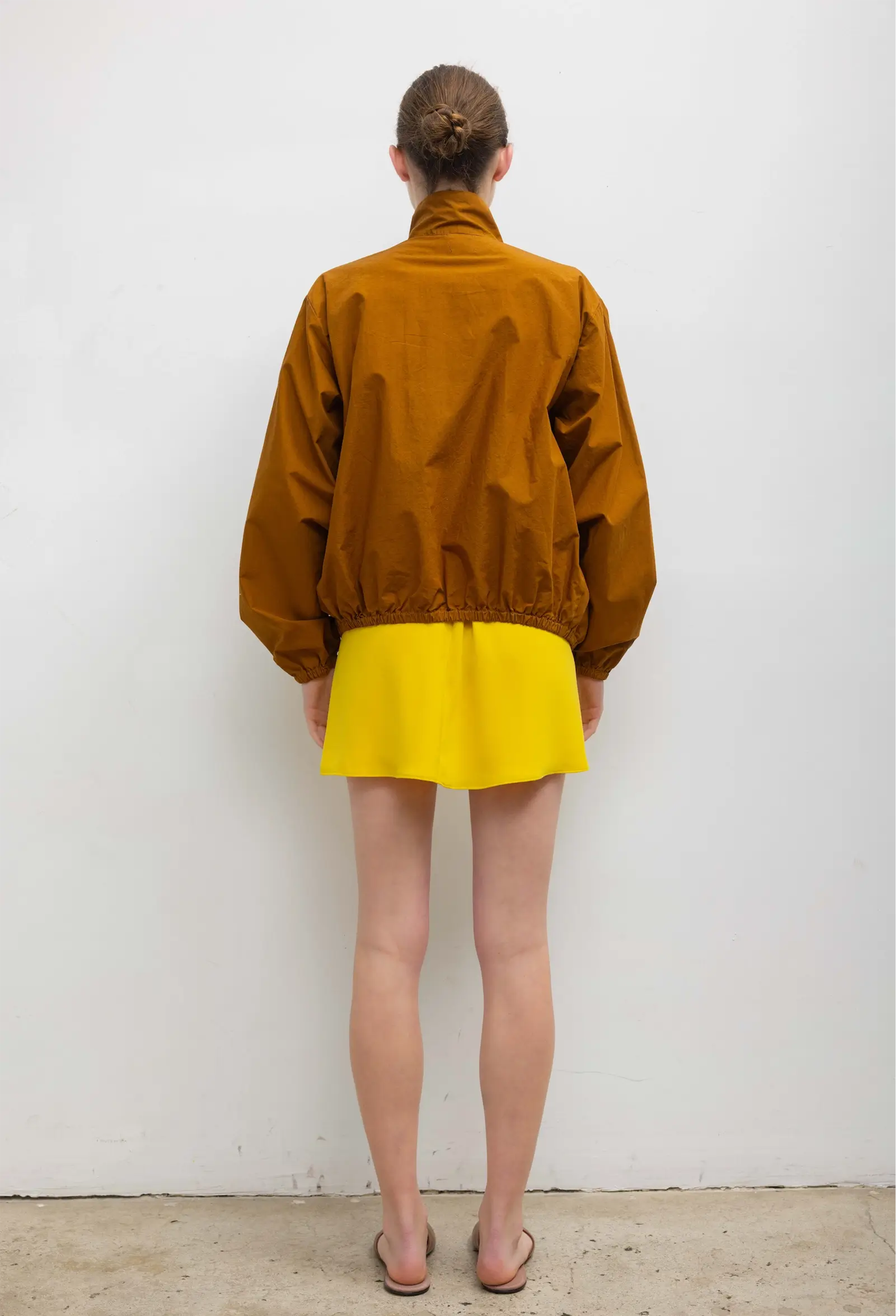 Person wearing a mustard jacket and yellow skirt against a white wall