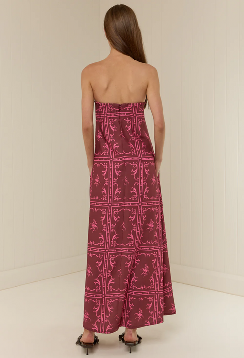 A woman viewed from behind wearing a strapless maxi dress with a magenta and dark red ornate pattern, paired with black pointed-toe heels, set against a plain off-white studio background.