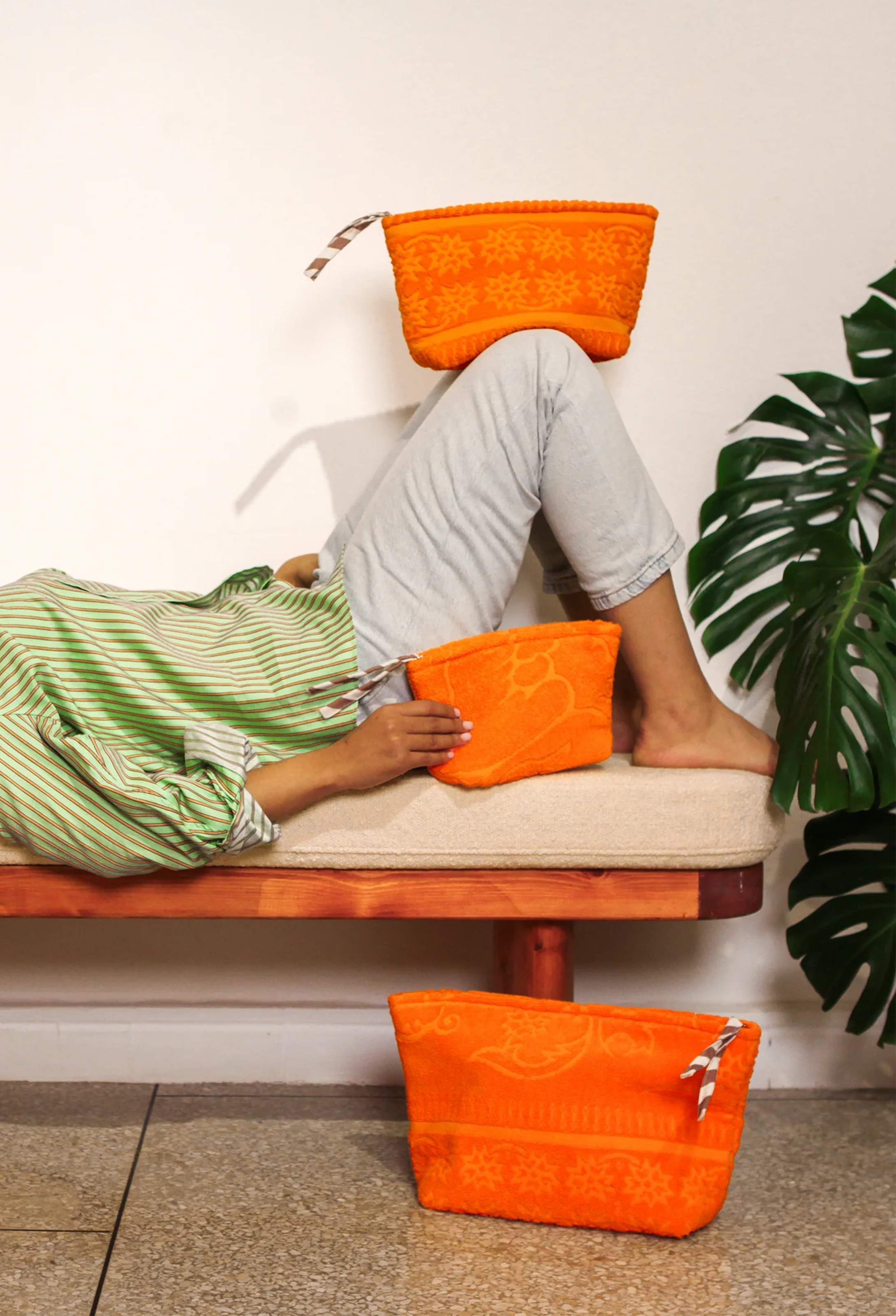 Three bright orange woven pouches with white zippers are arranged around a person lying on a cushioned bench in a styled indoor setting. a large green plant is visible at the right edge.