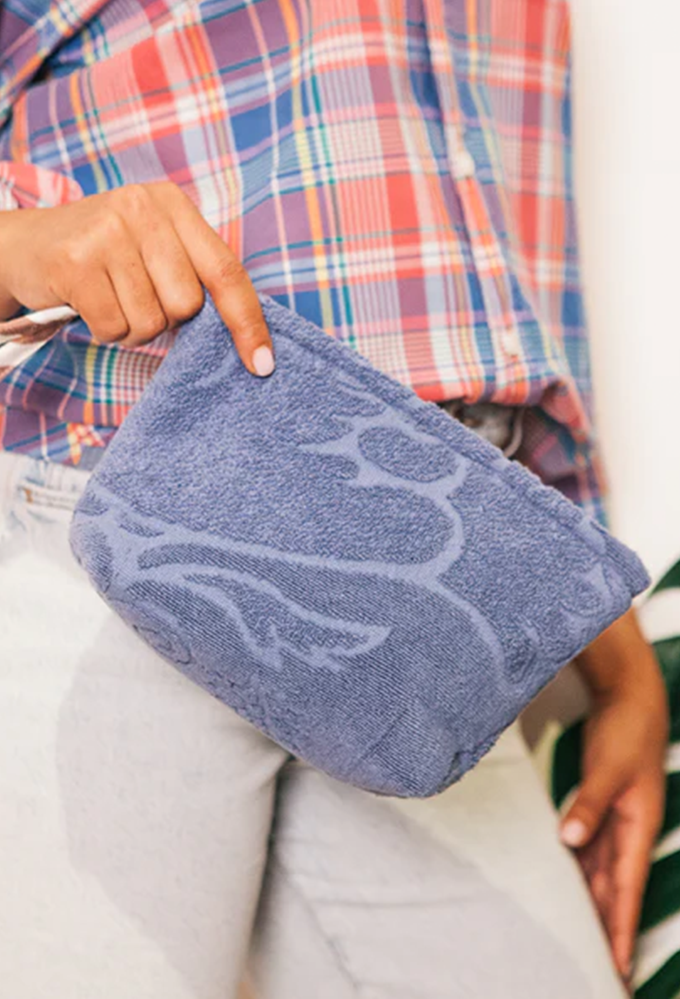 A person holding a dusty blue plush pouch with a white abstract pattern, wearing a red, blue, and white plaid shirt and light gray sweatpants.