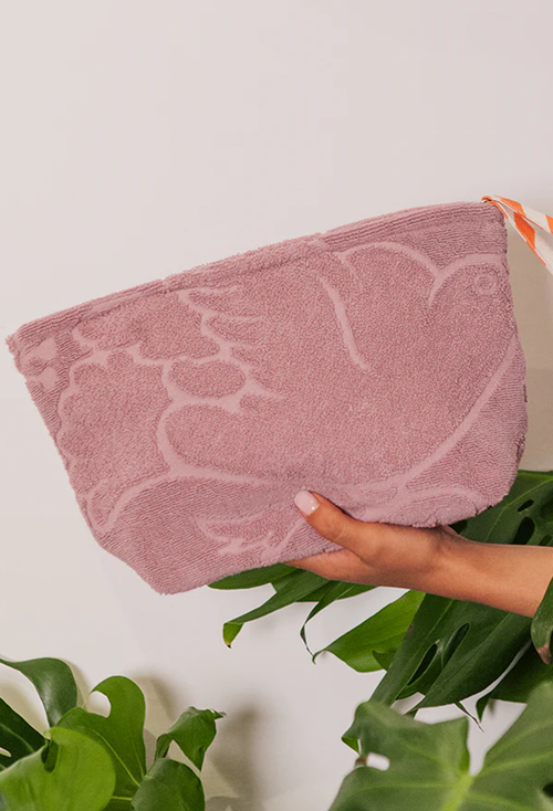 A dusty pink terrycloth pouch with a tonal  pattern, held by a hand against a white background with green plant leaves.