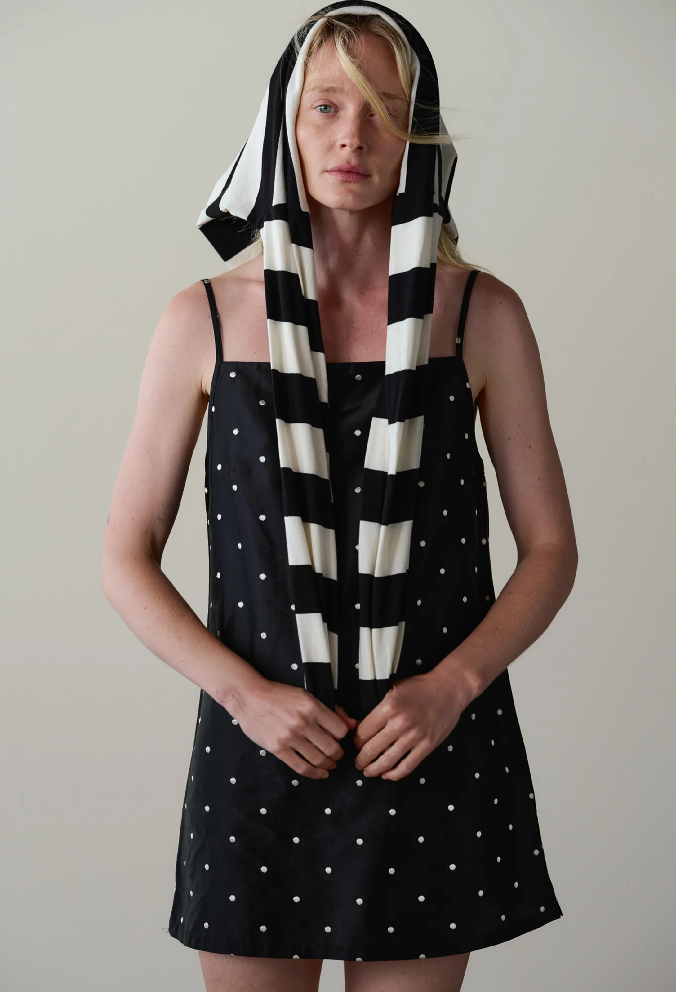 A model wearing a black and white striped and polka dot dress with a matching striped headscarf, standing against a plain light gray background.