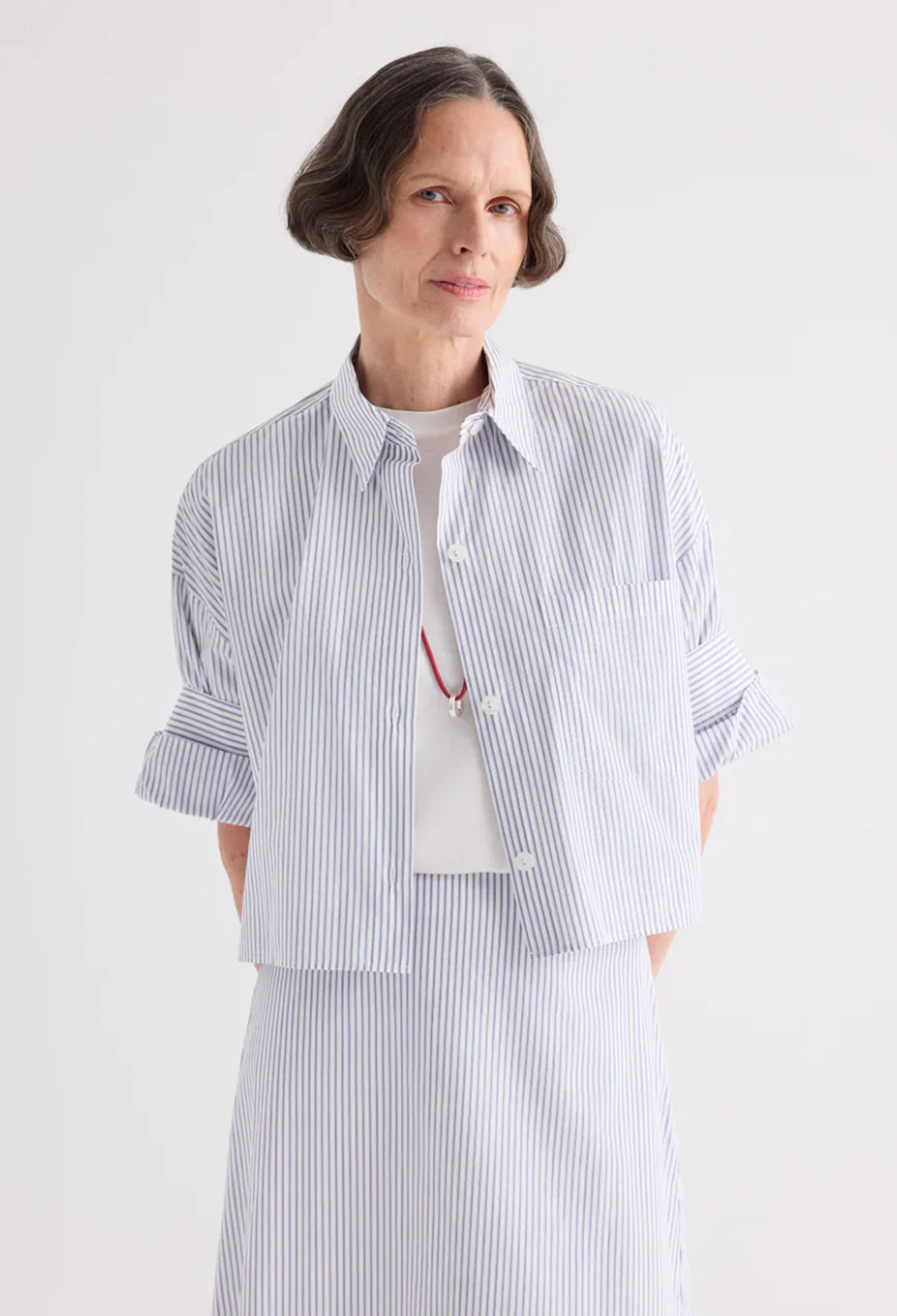 A model wearing a light blue and white vertically striped button-up shirt with rolled sleeves, layered over a white t-shirt and paired with a matching striped skirt, against a plain white studio background.
