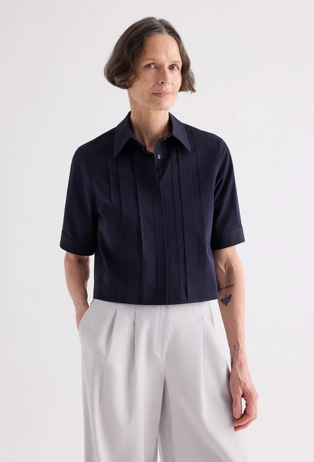 A woman modeling a short-sleeve dark navy pleated button-up shirt paired with light gray wide-leg trousers, photographed against a clean white studio background.