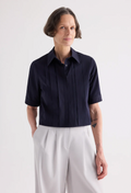 A woman modeling a short-sleeve dark navy pleated button-up shirt paired with light gray wide-leg trousers, photographed against a clean white studio background.