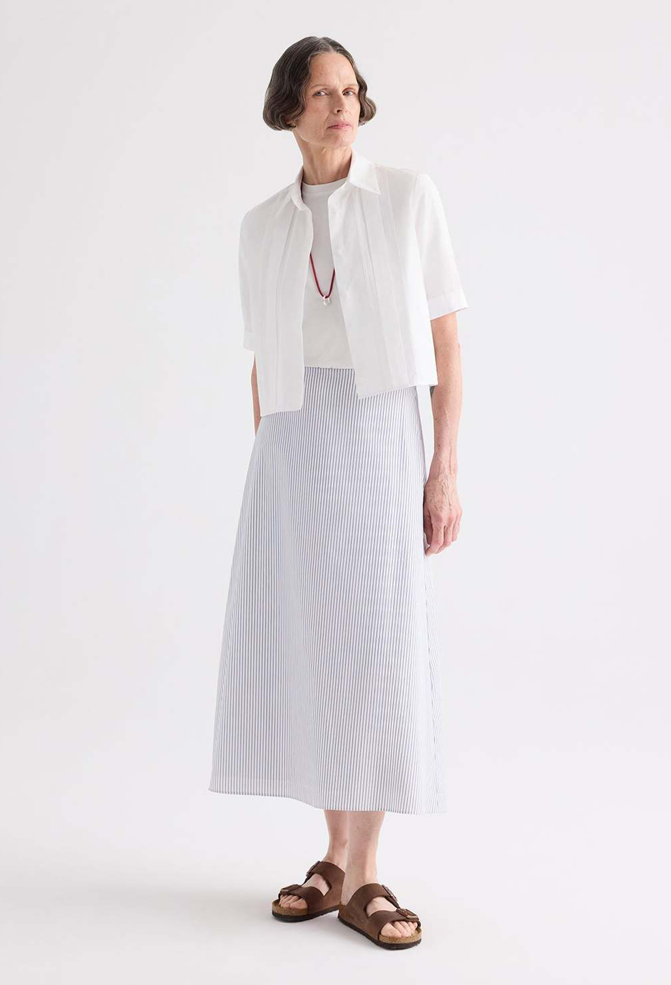 A model wearing a white short-sleeve cropped jacket over a white top and a white textured midi skirt, paired with brown sandals, against a plain white studio background.