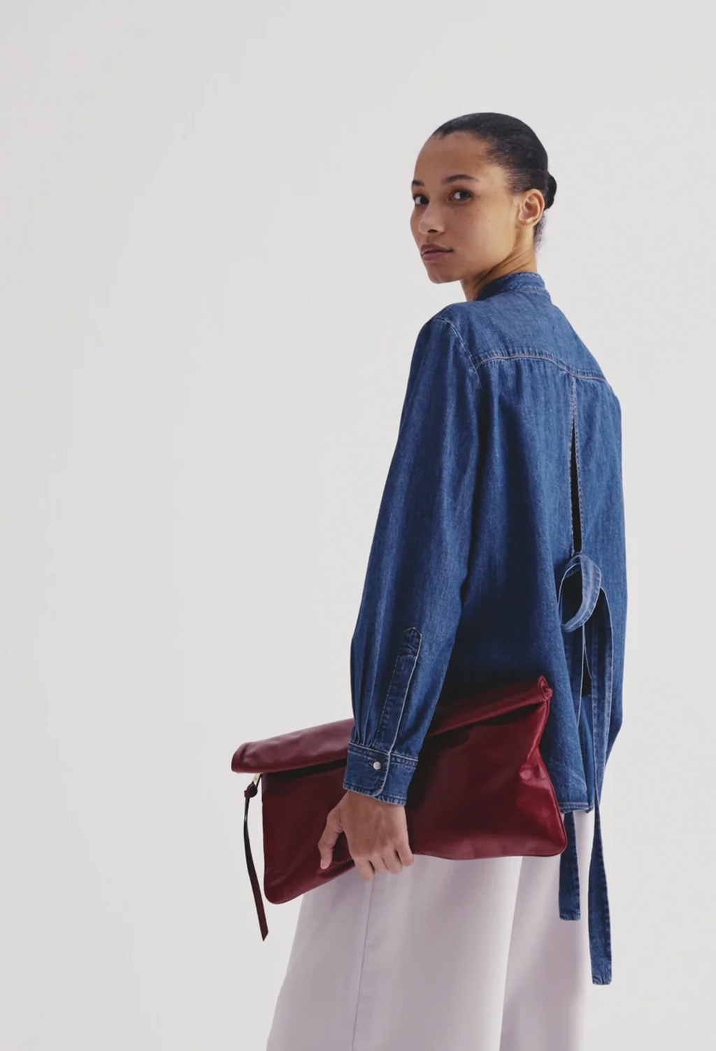 A model wearing a blue denim shirt and white trousers, holding a deep red leather clutch bag against a plain light gray background.