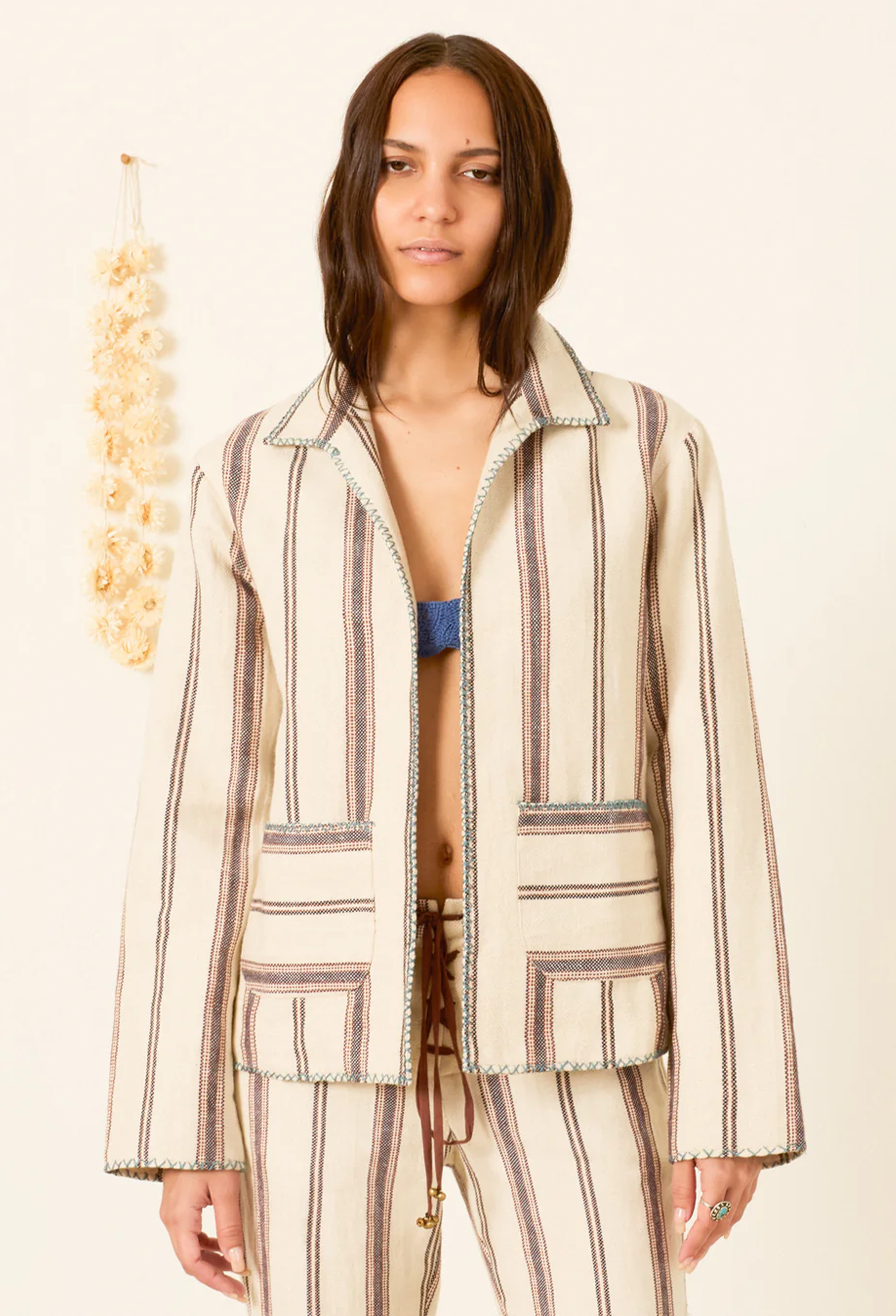 A woman modeling a matching striped jacket and trousers set in a cream color with brown and gray vertical stripes, styled with a blue bikini top against a warm off-white background.